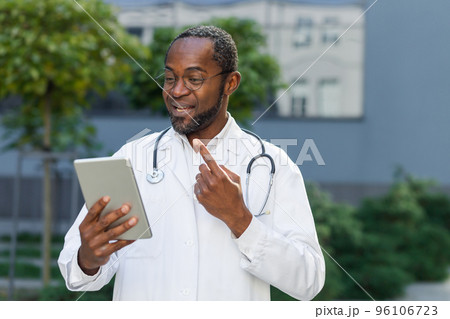 Smiling senior mature doctor in medical coat and glasses with tablet talking on video call remotely online, african american man outside clinic consulting patients. 96106723