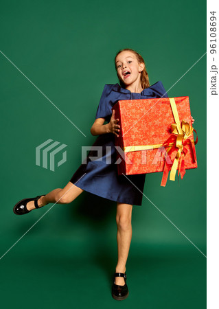 Portrait of little happy girl, child in dress posing with big present box isolated over green studio background 96108694