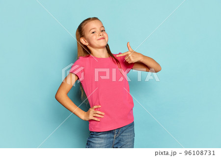 Portrait of little beautiful girl, child in pink T-shirt posing isolated over blue studio background. Proudly pointing at herself Portrait of little beautiful girl, child in pink T-shirt posing isolated over blue studio background. Proudly pointing at herself 96108731