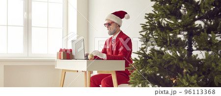Happy grey-haired bearded man in red suit, Santa cap sits at desk working on laptop near Xmas Tree. 96113368