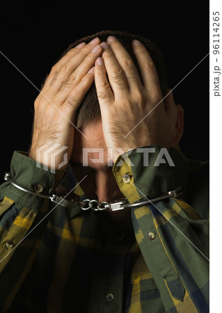 Portrait of a handcuffed man with his hands on his head. Caucasian man in green checkered shirt with cuffs on black background. Regret. Concept of crime and punishment. 96114265
