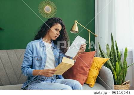 Young beautiful hispanic woman at home disappointed and sad received letter from university about bad exam results, student with curly hair sitting on sofa in living room. 96116543