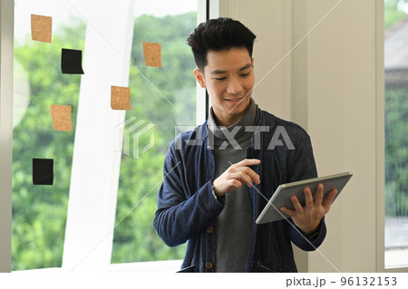Smiling young creative male freelancer using digital tablet while standing near window with sticky notes 96132153