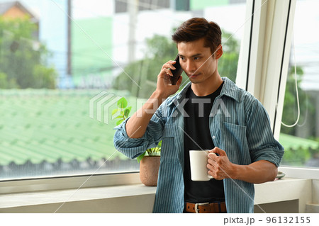 Handsome young man holding coffee cup and talking on mobile phone while standing near the office window 96132155