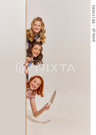 Portrait of three beautiful women peeking out corner with knives isolated over grey background. revenge 96133682