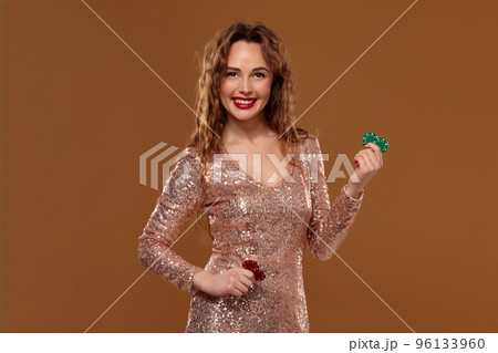 Blond woman weared in a gold glittered dress is holding some colorful gambling chips in her hands. Casino. 96133960