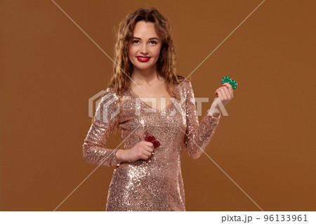Blond woman weared in a gold glittered dress is holding some colorful gambling chips in her hands. Casino. Blond woman weared in a gold glittered dress is holding some colorful gambling chips in her hands. Casino. 96133961