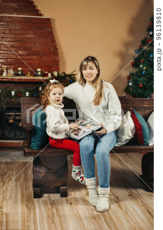 Cute mom and daughter are looking through a family photo album on Christmas Eve, spending time together by the fireplace and the Christmas tree. Christmas, New Year, memories, family evening 96139810
