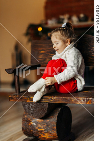 A charming baby is sitting on a bench against the background of the fireplace and putting on warm socks. The concept of Christmas and New Year, on the eve of holidays 96139815
