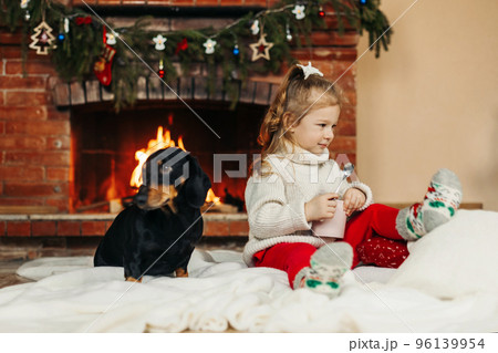 A happy little girl is playing with her dog at home, drinking hot chocolate on Christmas Eve A happy little girl is playing with her dog at home, drinking hot chocolate on Christmas Eve 96139954