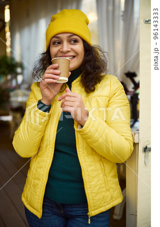 Waist-up portrait of multi-ethnic charming woman, dressed in green warm sweater, yellow wool hat and jacket, smiling, looking aside while enjoying her coffee break, taking a sip of hot aroma coffee Waist-up portrait of multi-ethnic charming woman, dressed in green warm sweater, yellow wool hat and jacket, smiling, looking aside while enjoying her coffee break, taking a sip of hot aroma coffee 96143333