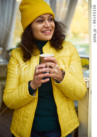 Close-up portrait of an attractive brunette multi-ethnic woman, in bright yellow hat and jacket, smiling a cheerful tothy smile looking aside, warming her hands with a takeaway hot coffee in paper cup 96143336
