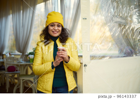 Attractive curly woman, wearing a green jacket, bright yellow hat and jacket, warming her hands with a takeaway hot aroma coffee in a disposable paper cup, smiling a beautiful toothy smile to camera Attractive curly woman, wearing a green jacket, bright yellow hat and jacket, warming her hands with a takeaway hot aroma coffee in a disposable paper cup, smiling a beautiful toothy smile to camera 96143337