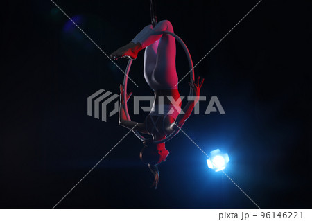 Young woman performing acrobatic element on aerial ring against dark background Young woman performing acrobatic element on aerial ring against dark background 96146221