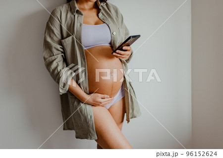 Pregnant woman using phone app for online pregnancy prenatal class or reading about motherhood wearing underwear and casual cotton shirt at home. Relaxing holding expecting belly for health. 96152624