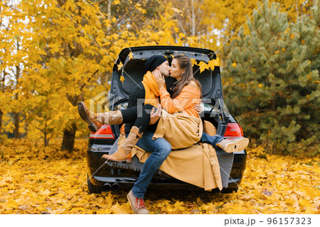 A cute young couple in love is sitting in a car with the trunk open, hugging, ready to kiss the autumn forest for the first time. The concept of travel and love A cute young couple in love is sitting in a car with the trunk open, hugging, ready to kiss the autumn forest for the first time. The concept of travel and love 96157323