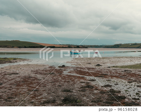 Clonakilty Bay at low tide on a day. A small blue fishing boat is anchored. Open seabed, silt and algae. Picturesque seaside landscape. 96158028