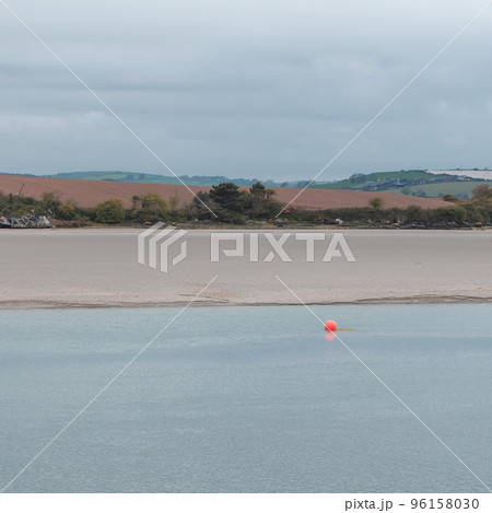 A small orange buoy is kept on a calm water surface. The sandy shore of the sea bay. The coastline on a cloudy day. Seaside landscape. 96158030