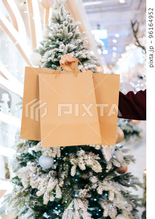 Close-up vertical cropped shot of unrecognizable man holding in hand paper bags with gifts in hall of celebrate shopping mall in Christmas eve, on background of bright beautiful xmas decorations. Close-up vertical cropped shot of unrecognizable man holding in hand paper bags with gifts in hall of celebrate shopping mall in Christmas eve, on background of bright beautiful xmas decorations. 96164952