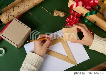 Top view woman's hands, holds cardboard box, calculates the amount of wrapping material for packing gifts for Christmas, Women's, Saint Valentine's Day or any other event using wooden ruler and pencil Top view woman's hands, holds cardboard box, calculates the amount of wrapping material for packing gifts for Christmas, Women's, Saint Valentine's Day or any other event using wooden ruler and pencil 96168225