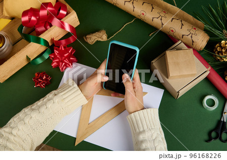 Selective focus on woman's hands holding a smartphone empty blank touch screen, with copy ad space for promotional text, over a green background with Xmas wrapping materials, decorative tapes and bows Selective focus on woman's hands holding a smartphone empty blank touch screen, with copy ad space for promotional text, over a green background with Xmas wrapping materials, decorative tapes and bows 96168226
