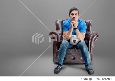 Asian man with the ball sitting on the couch with a serious expression Asian man with the ball sitting on the couch with a serious expression 96169201