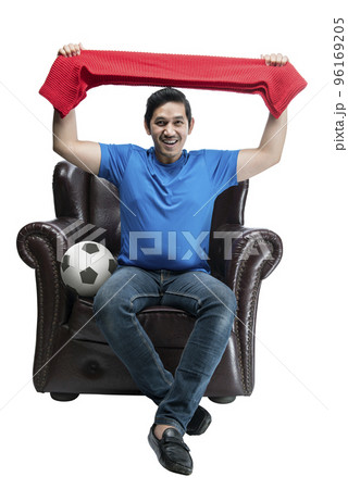 Asian man with the ball holding scarf sitting on the couch with an excited expression Asian man with the ball holding scarf sitting on the couch with an excited expression 96169205