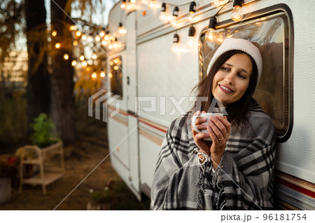 Caucasian woman in a knitted hat wrapped in a plaid and drinks a warming drink outdoors. Travel in a motor home in the fall. 96181754