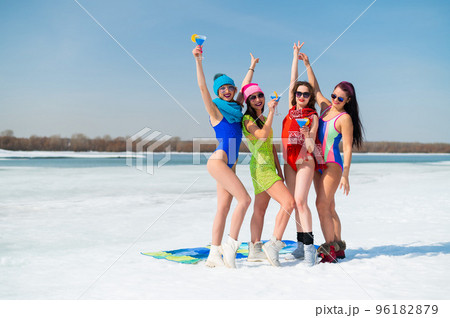 Four caucasian women in swimsuits pose and drink blue cocktails on a snowy beach. 96182879