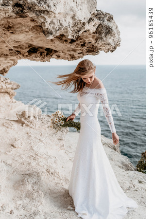 Romantic bride, blond girl in white wedding dress with open shoulders posing with sea and rocks in background. Stylish young woman standing on cliff's edge and looking into a ocean mountains at sunset 96185439