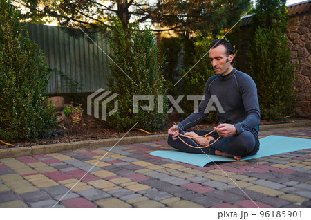Peaceful man yogi sitting in lotus pose on a fitness mat, meditating with rosary beads outdoors at sunset. Yoga practice. Meditation. Enlightenment. Body conscious. Active and healthy lifestyle 96185901