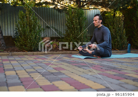 Peaceful middle-aged Caucasian man, a yogi sitting on a fitness mat, meditating with rosary beads while practicing yoga outdoors. Mindfulness. Spiritual growth. Awakening. Enlightenment. Meditation 96185904