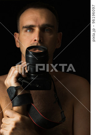 Close-up of a man with a camera against a black background. Photographer holding camera at mouth level. Breast portrait of a Caucasian man with a DSLR camera shirtless. Low key. Close-up of a man with a camera against a black background. Photographer holding camera at mouth level. Breast portrait of a Caucasian man with a DSLR camera shirtless. Low key. 96196097