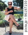 A middle-aged sexy woman in a summer skirt and T-shirt slightly crossed bare legs, and sunglasses sits on a bench in the park with a pensive look and looks at her hand 96197150