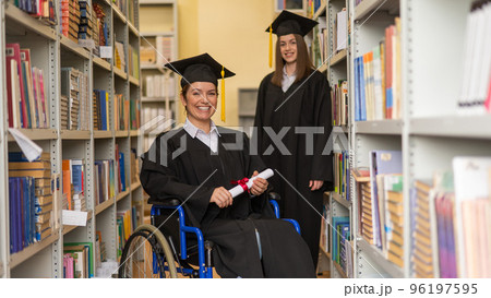 Happy young woman and woman in wheelchair in graduate gown with diploma in hands in library. Inclusive education. 96197595