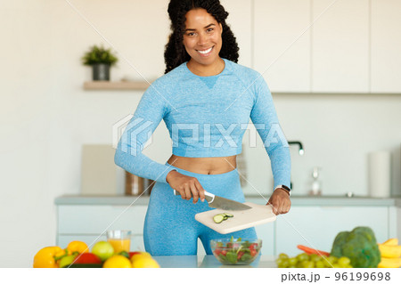 Happy fit black woman in sportswear preparing fresh vegetable salad, standing in the kitchen after domestic workout 96199698