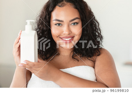 Beautiful black plus size woman holding bottle with moisturising body lotion, using new skincare product after bath 96199810