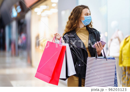 Young woman in protective mask using mobile phone while holding shopping bag in department store. Young woman in protective mask using mobile phone while holding shopping bag in department store. 96203123