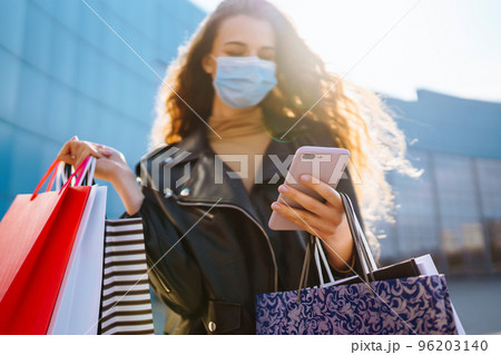 Smiling woman in protective medical mask with shopping bags using her phone. Young woman after. Smiling woman in protective medical mask with shopping bags using her phone. Young woman after. 96203140