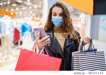 Young woman in protective mask using mobile phone while holding shopping bag in department store. 96203143