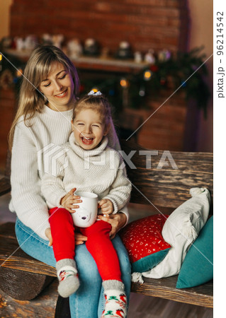 Portrait of a happy little girl sitting on her mother's lap and drinking hot chocolate on Christmas or New Year's Eve. Christmas mood, Christmas spirit, time together, motherhood, childhood 96214542