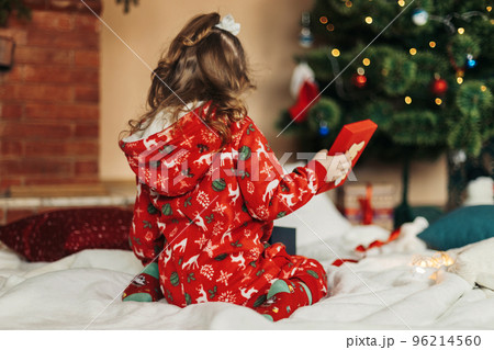 A little girl opens a Christmas present near the Christmas tree. Christmas time, gifts, festive mood. The concept of Christmas and New Year. Rear view A little girl opens a Christmas present near the Christmas tree. Christmas time, gifts, festive mood. The concept of Christmas and New Year. Rear view 96214560