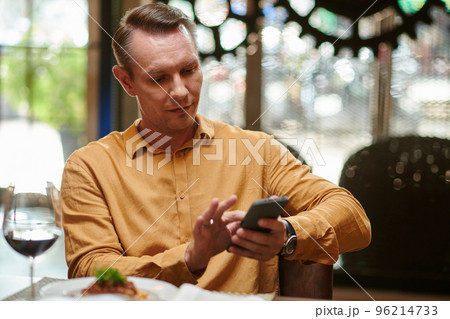 Smiling man sitting at table in restaurant and checking social media on smartphone Smiling man sitting at table in restaurant and checking social media on smartphone 96214733