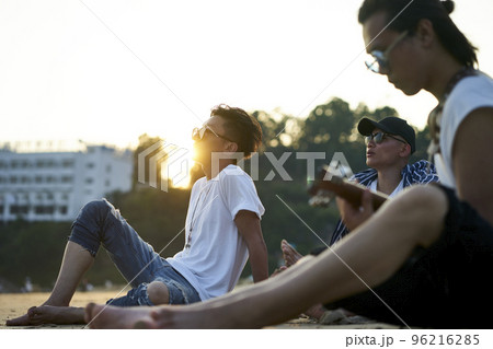 three young asian adult men sitting on sand beach playing guitar at dusk three young asian adult men sitting on sand beach playing guitar at dusk 96216285