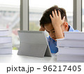 Young Asian elementary school boy sitting next to stack of books and using tablet computer with both hands holding his head with gestures and face showing stress. 96217405