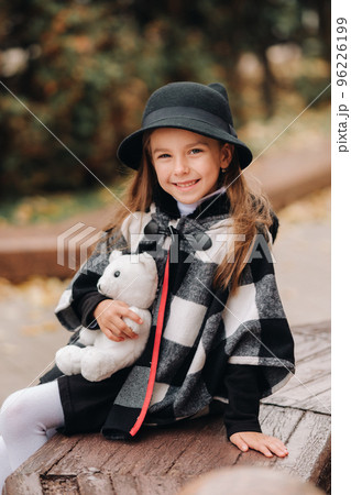 A little stylish girl in autumn clothes is sitting on a bench in the autumn city 96226199