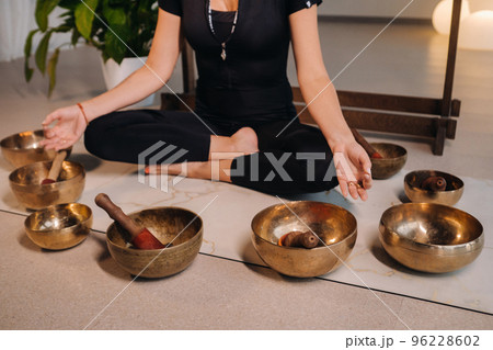 A woman sits in a lotus position next to Tibetan bowls, sitting on a yoga mat against the background of a gong 96228602