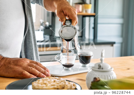 Man preparing classic Italian coffee in the mocha in the kitchen. Pouring coffee from moka pot into small glass coffee cup. Coffee brake. Morning habit. 96228744