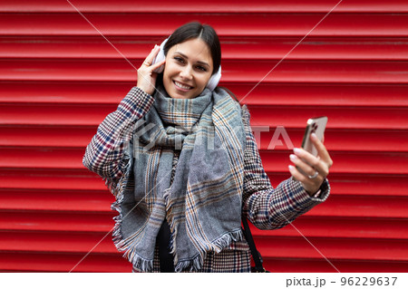 stylish adult woman in headphones with a mobile phone in her hands listens to music against a red stylish adult woman in headphones with a mobile phone in her hands listens to music against a red 96229637