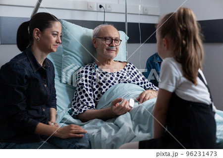 Elderly man lying in hospital bed chatting with visiting family members. Little girl talking to grandfather in elderly people medical clinic. Family living with old man resting in sanatorium bed. 96231473